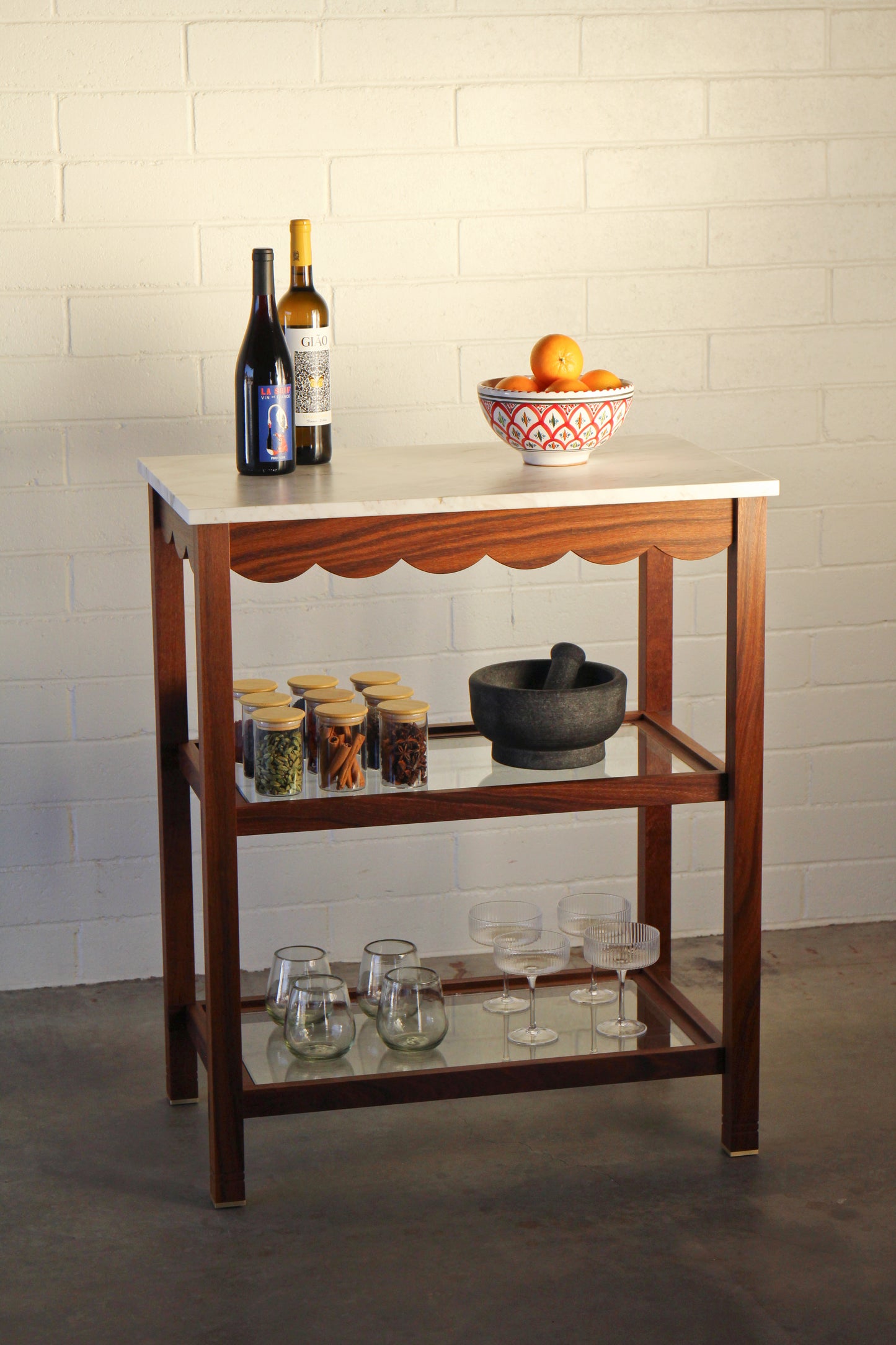 Wooden bar cart with bottles, a bowl, and kitchen items against a white brick wall.
